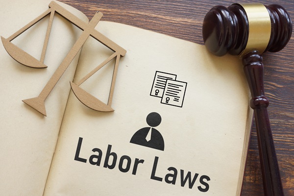 Open book displaying the words "Labor Laws" with icons of legal documents and a person, next to a wooden gavel and scale symbol on a wooden table.