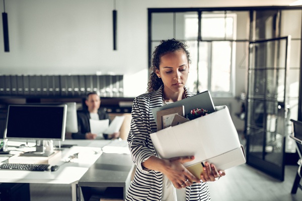 Woman carrying a box of personal belongings while leaving an office, representing job termination or potential wrongful dismissal.
