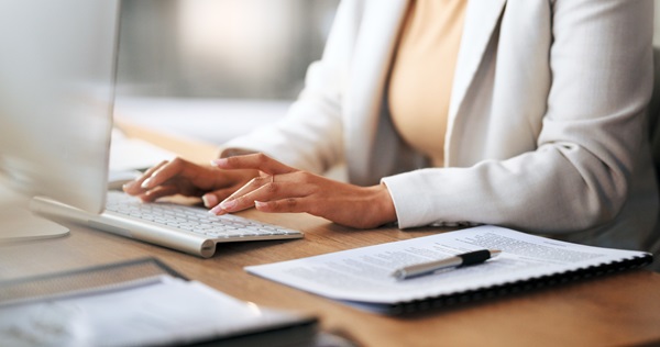 A close-up of a person in a white blazer typing on a computer keyboard at a desk.