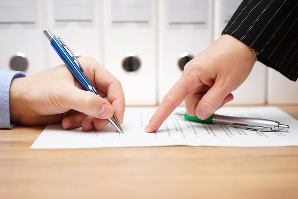 A close-up shot of a person’s hand holding a blue pen, prepared to sign a severance agreement on a wooden desk.